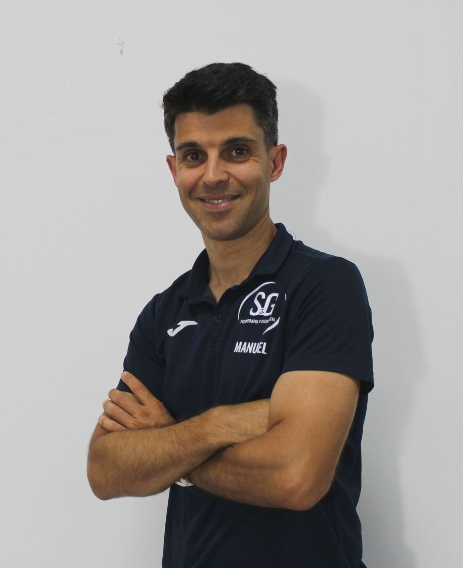 Hombre con los brazos cruzados, vistiendo una camisa azul oscuro con logo, sonriendo sobre un fondo blanco.