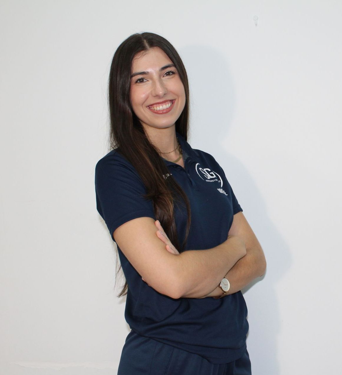 Mujer de cabello largo y oscuro, con un polo azul marino con el logotipo, con los brazos cruzados y sonriendo. Fondo blanco.