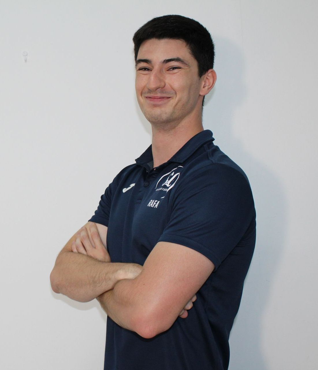 Hombre con los brazos cruzados, sonriendo. Vistiendo un polo azul marino sobre un fondo blanco liso.