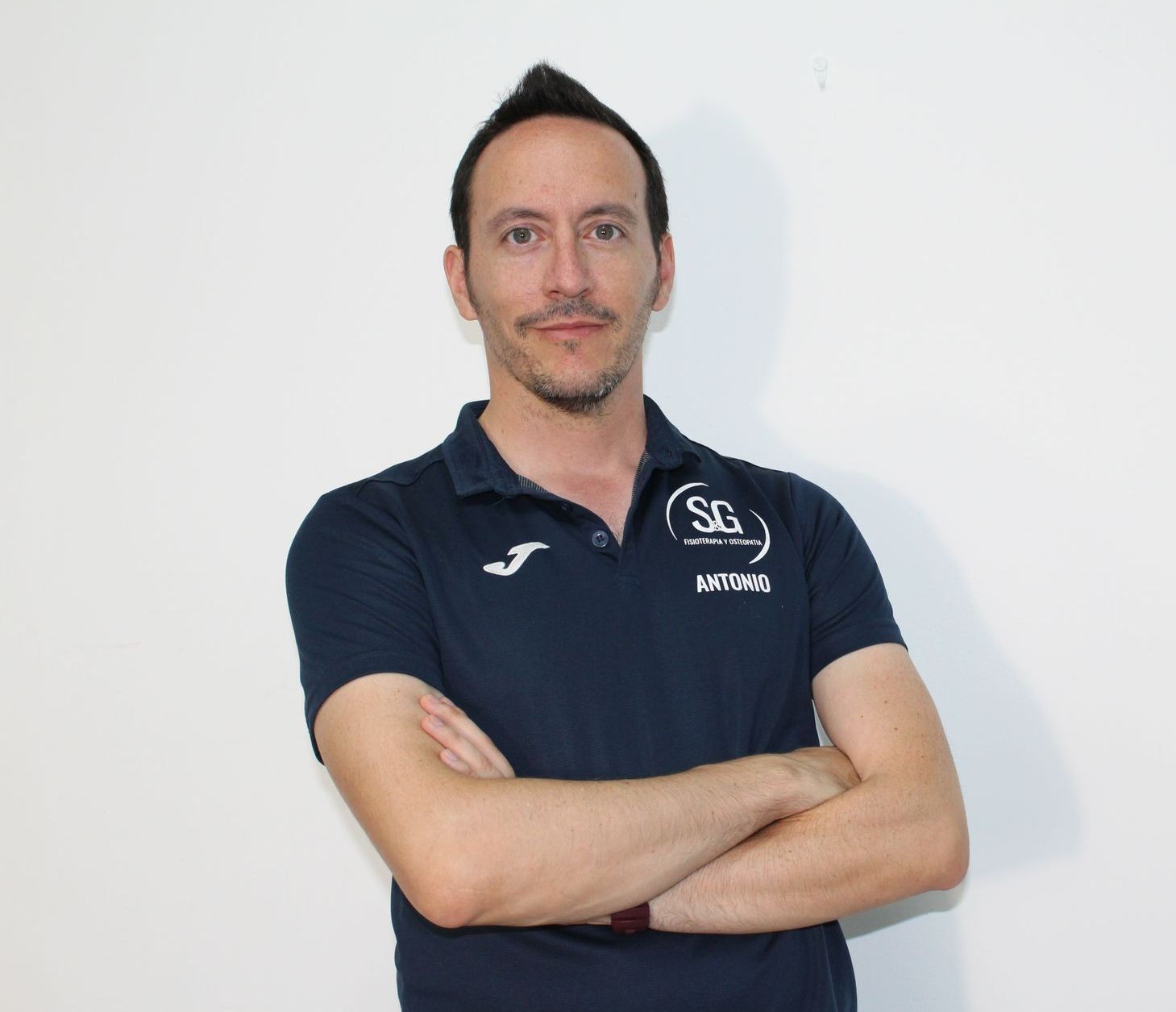 Hombre con camisa azul oscuro y brazos cruzados, de pie frente a una pared blanca.