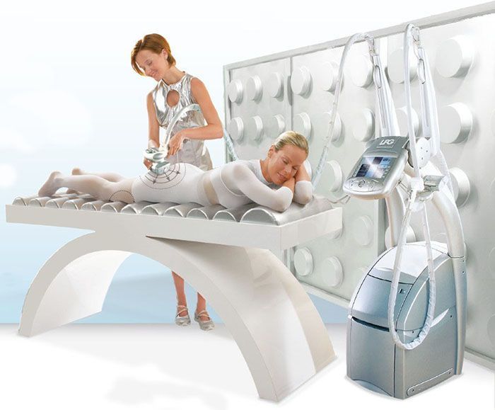 Mujer recibiendo tratamiento corporal en un spa; un técnico usa un dispositivo. Habitación blanca, cama y máquina.