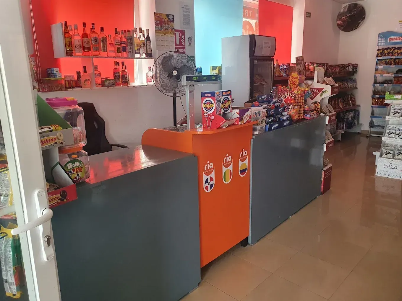Mostrador de una pequeña tienda con gabinetes de color naranja y gris oscuro, estantes con artículos y piso de baldosas.