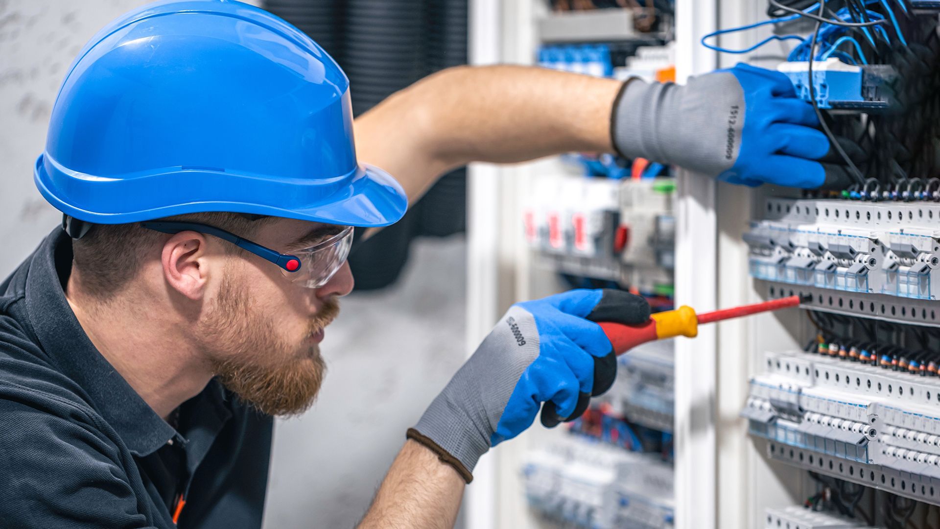Electricista con casco azul y gafas de seguridad trabajando en un panel, utilizando un destornillador.