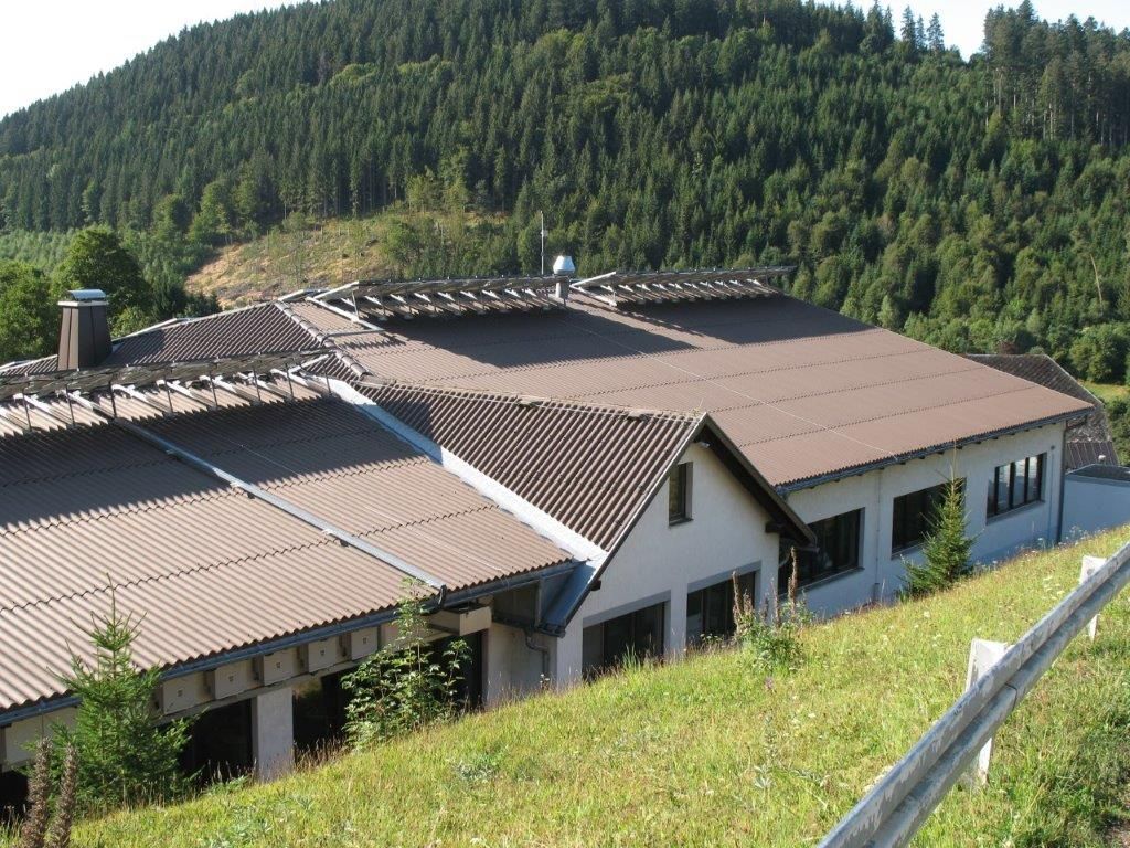 Flaches Gebäude mit braunem Welledach inmitten einer bergigen Waldlandschaft
