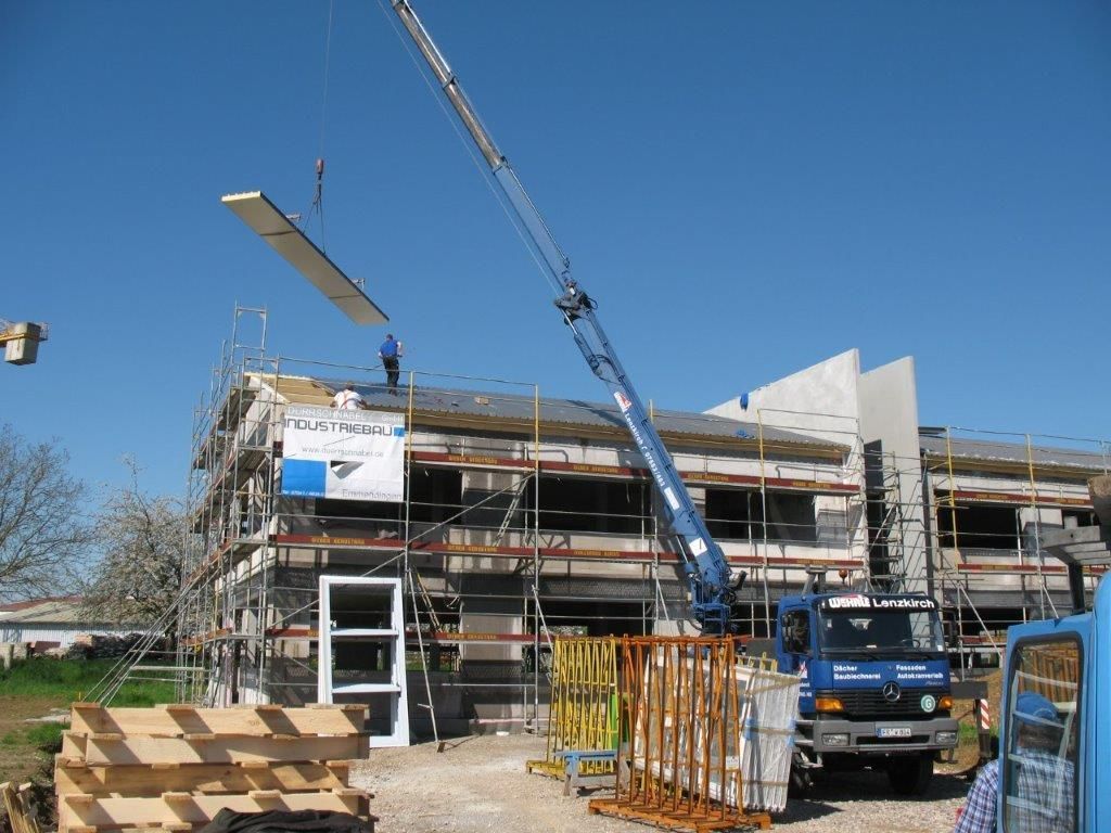 Ein Kran hievt ein langes Bauteil auf ein im Bau befindliches Holzhaus