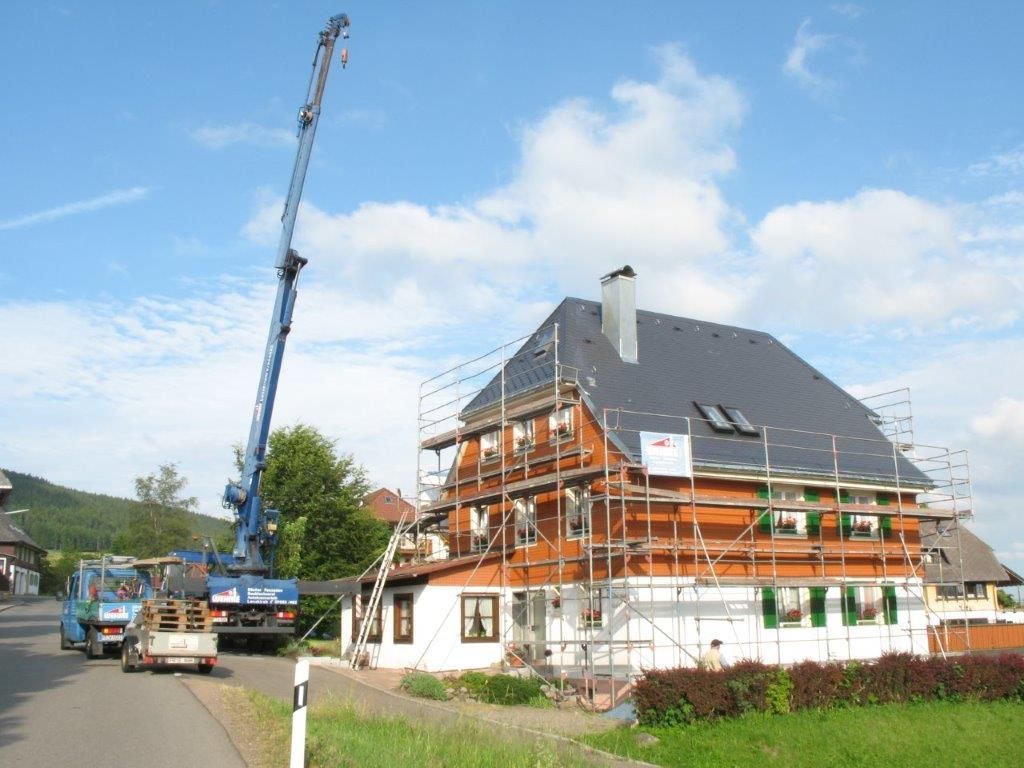 Ein Mobilkran hebt Baumaterial über ein traditionelles Holzhaus vor bewaldetem Hügel