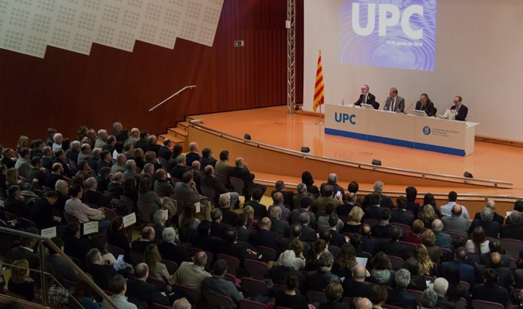 Personas sentadas en un auditorio durante un evento de la UPC, frente a un escenario con cuatro oradores.