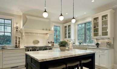 Cocina luminosa con una isla central oscura, armarios blancos, encimeras de mármol y tres lámparas colgantes.