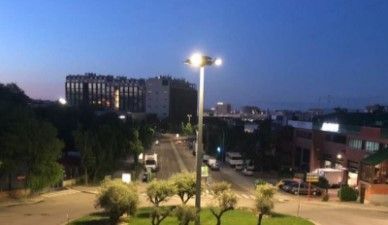 Una escena callejera al anochecer con una farola brillante, vegetación verde, coches aparcados y edificios al fondo.