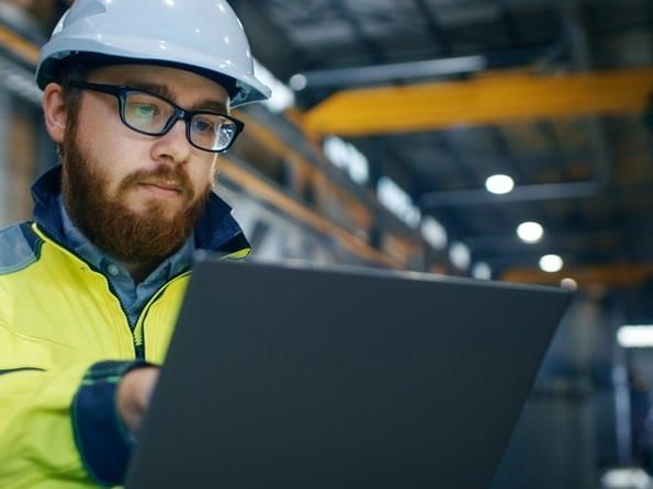 Una persona que lleva un casco blanco y un chaleco amarillo de alta visibilidad utiliza un ordenador portáti.