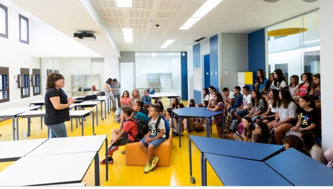 Una profesora se dirige a un numeroso grupo de alumnos sentados en un aula luminosa y moderna con suelo.