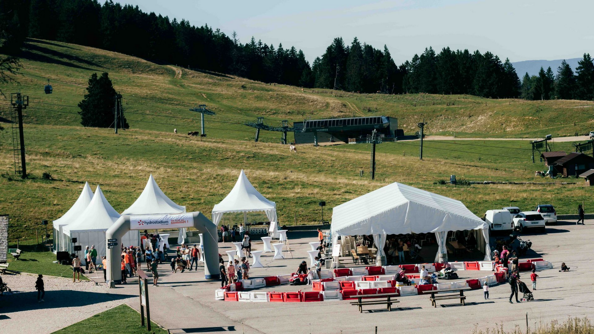 Événement en plein air avec des tentes blanches, des gens et une colline herbeuse avec un télésiège.