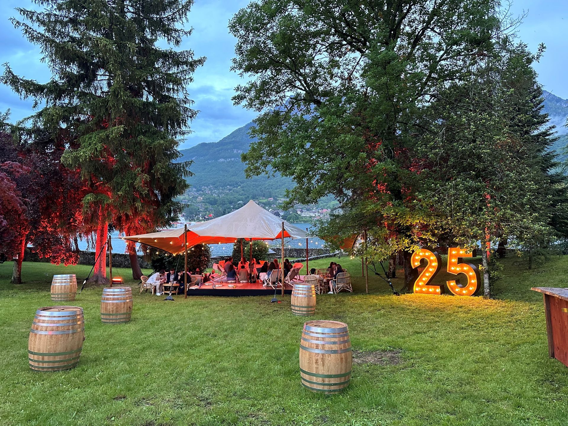 Une fête en plein air avec une tente, des barils et une enseigne lumineuse 25. Des gens sont rassemblés ; arbres, lac et montagnes en arrière-plan.