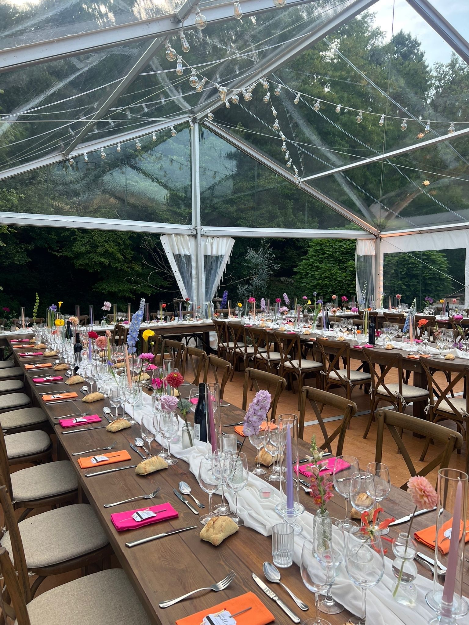 De longues tables en bois sont dressées pour une fête sous une tente transparente, avec des centres de table floraux et des chaises.