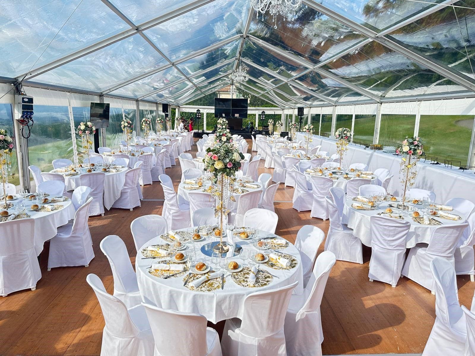 Aménagement de la réception de mariage : tables rondes avec nappes blanches, chaises, centres de table floraux sous une tente transparente.