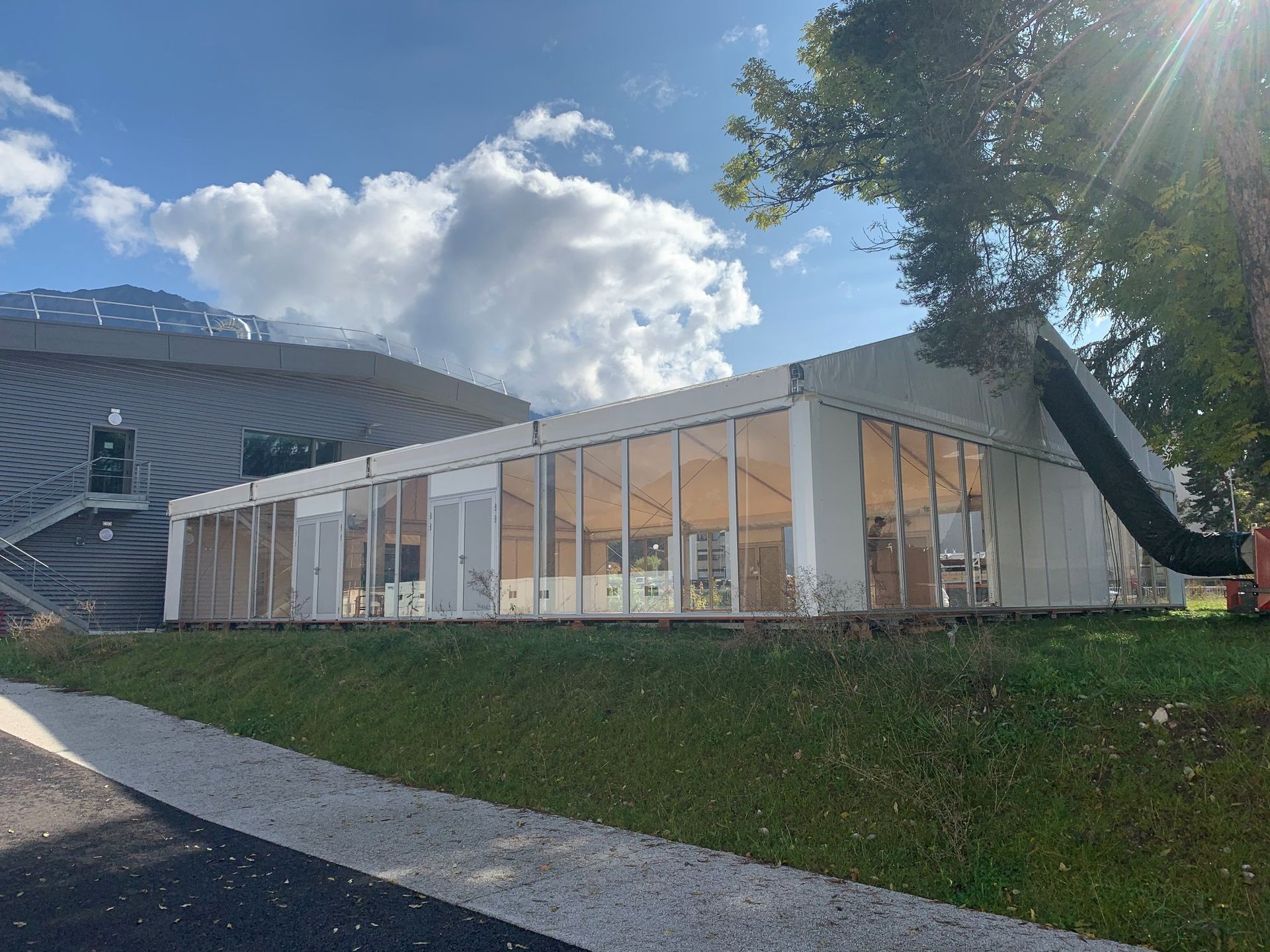 Tente événementielle blanche avec fenêtres transparentes, à côté d'un bâtiment et d'une zone herbeuse sous un ciel nuageux.