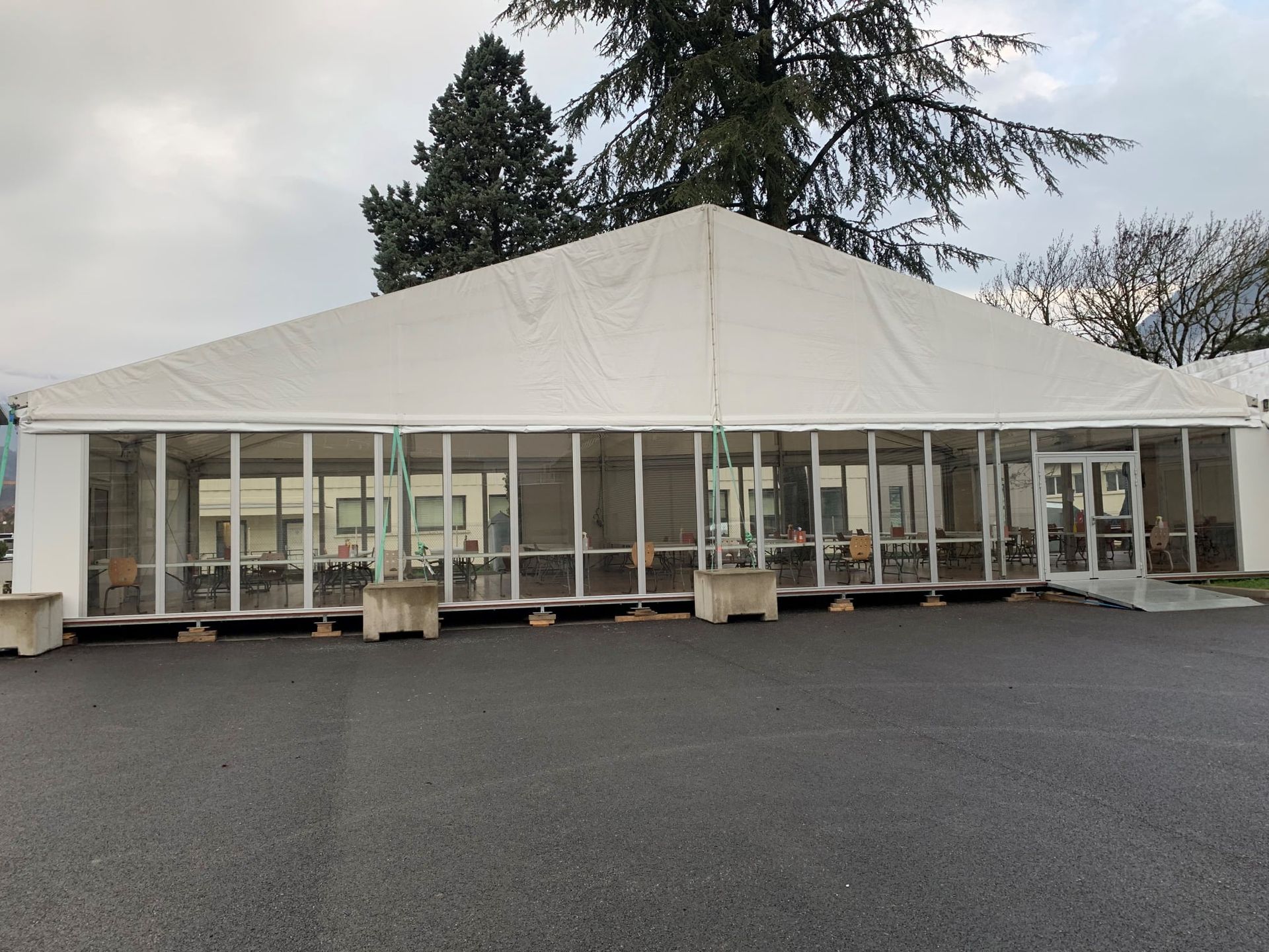 Tente blanche aux parois de verre sur asphalte noir, arbres en arrière-plan.