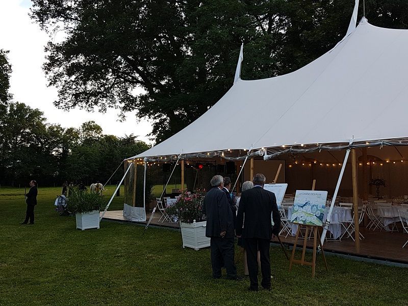 Une tente blanche illuminée de guirlandes lumineuses est dressée sur une pelouse ; des gens se rassemblent autour d'œuvres d'art.