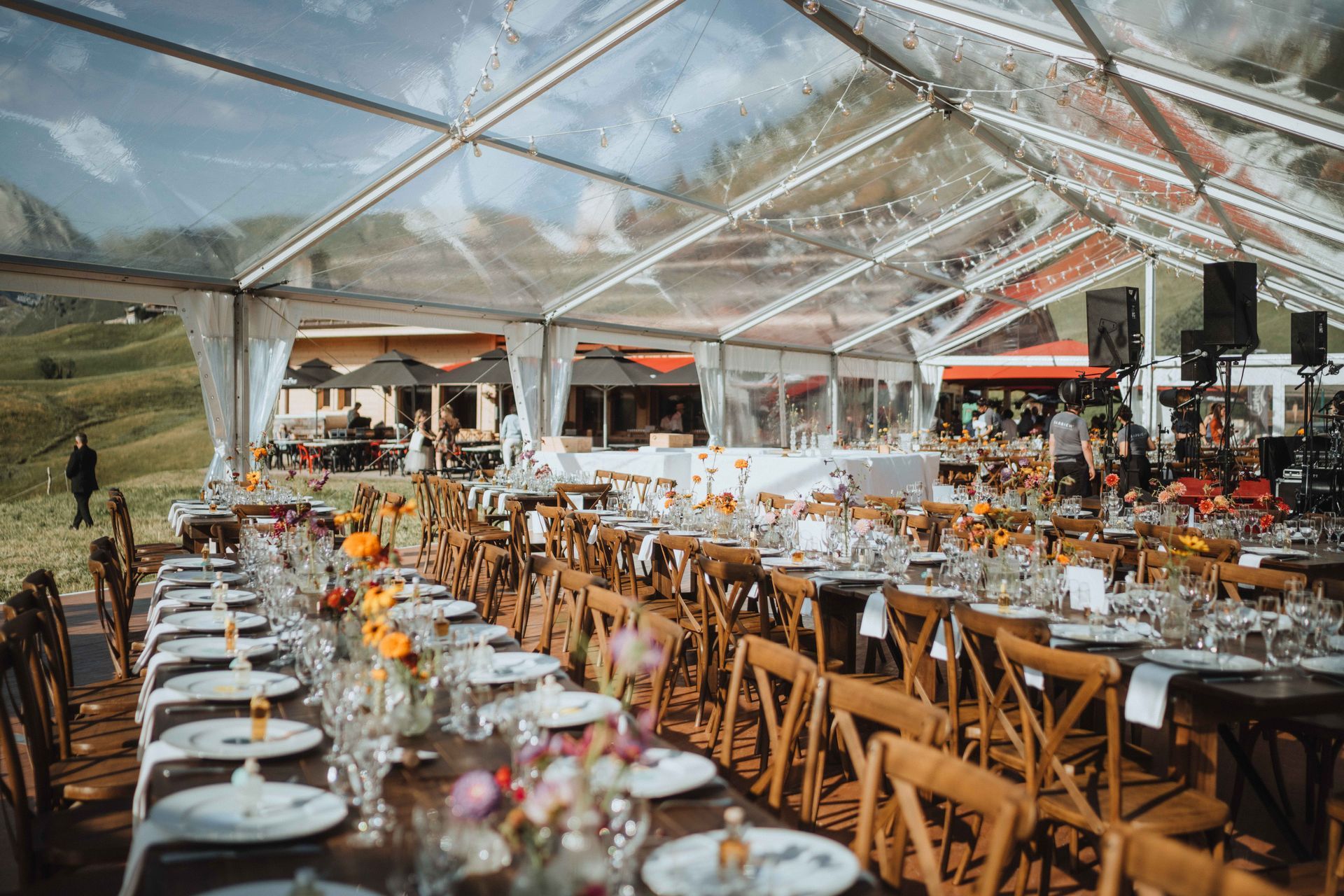 De longues tables sont dressées pour une réception de mariage sous une tente transparente, avec des compositions florales et un vignoble en arrière-plan.