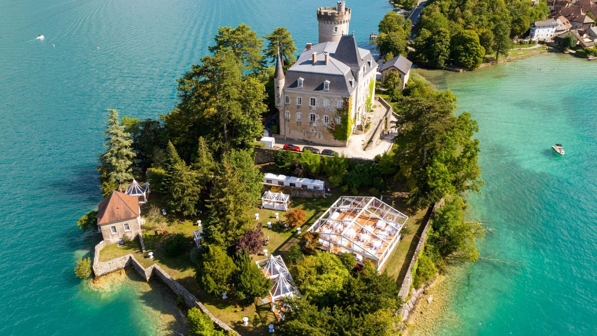 Vue aérienne : Une longue tente transparente est installée à flanc de colline, attenante à un château. Des arbres entourent le site.