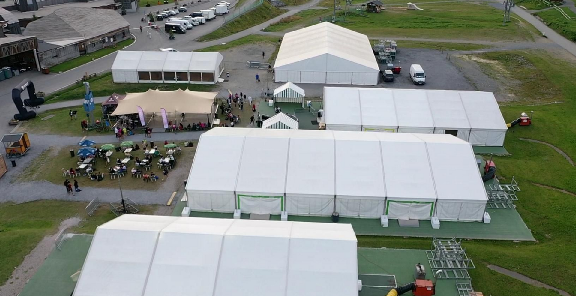 Vue aérienne d'un événement en plein air avec de nombreuses tentes blanches, des personnes et des véhicules sur une zone herbeuse.