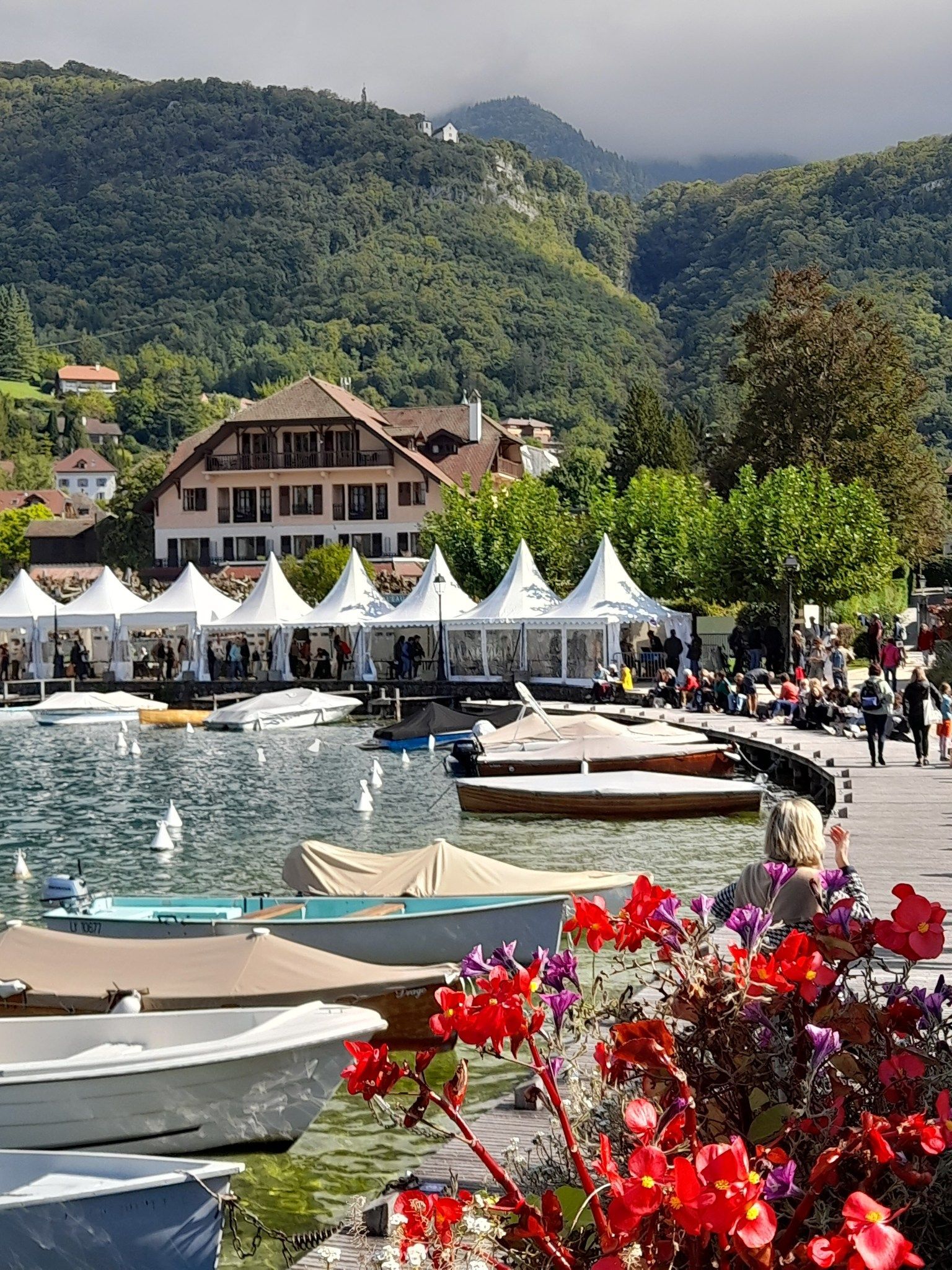 Bateaux sur le lac, tentes blanches, personnes, bâtiments, montagnes en arrière-plan, fleurs rouges au premier plan.