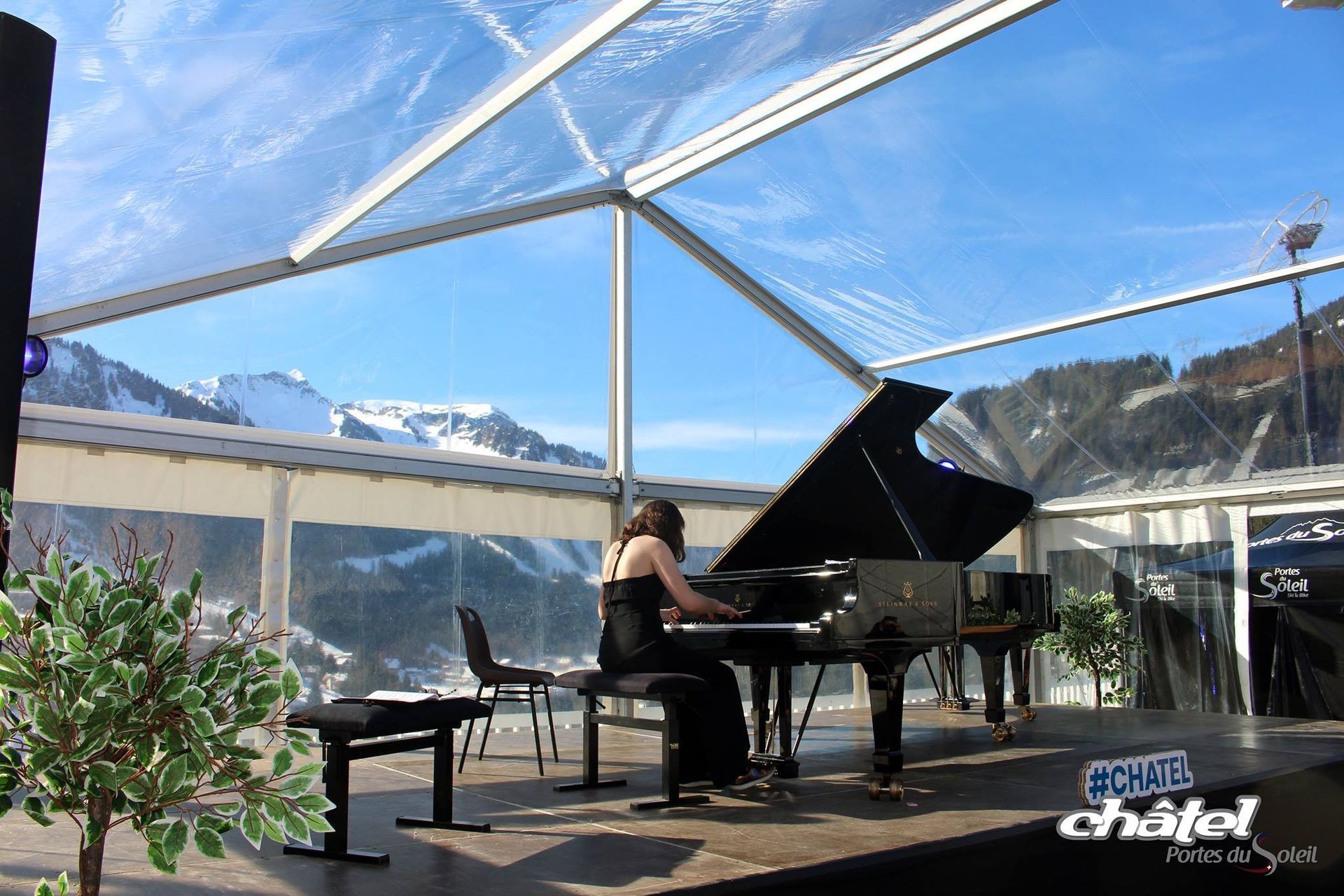 Un pianiste joue du piano à queue sous une tente, les montagnes sont visibles à travers les parois transparentes.