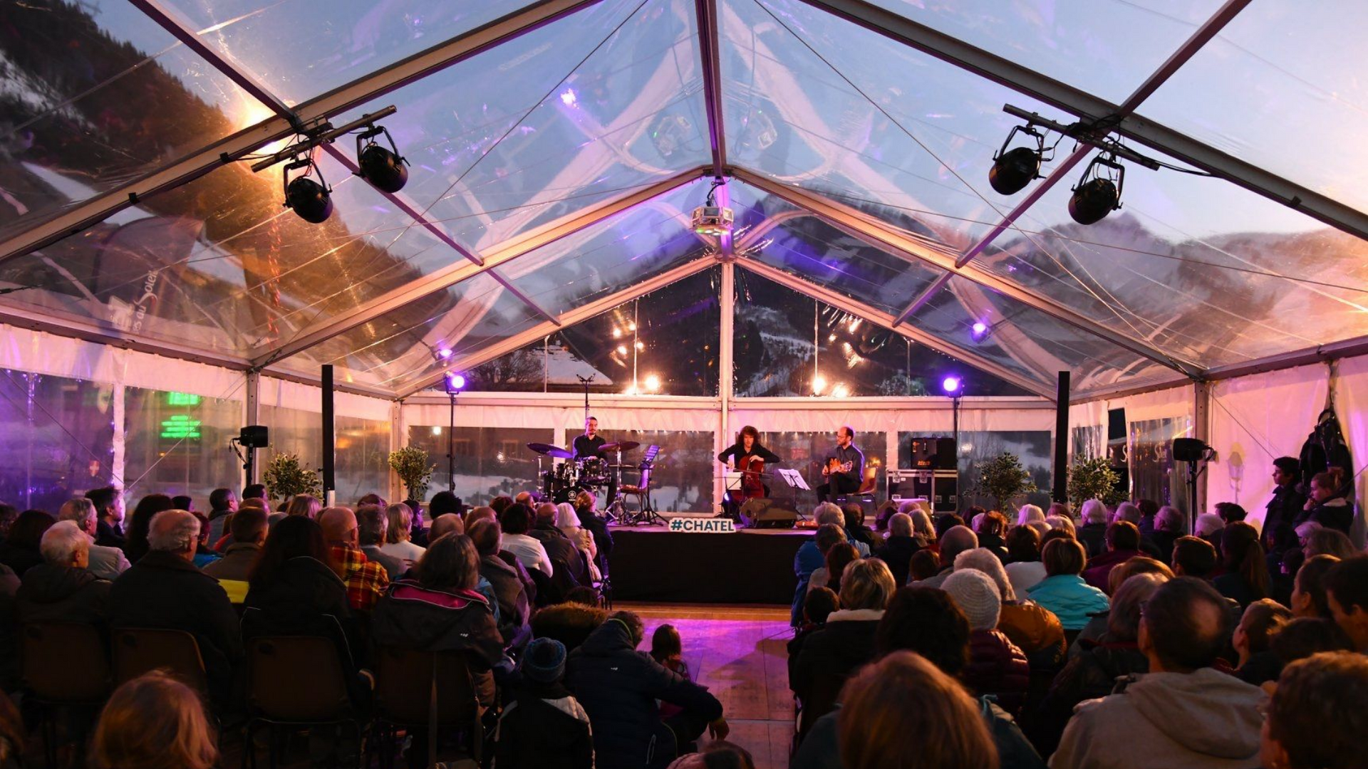 Un groupe joue sur scène sous un chapiteau transparent, le public est assis. Éclairage crépusculaire avec les montagnes en arrière-plan.