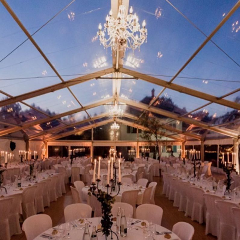 Tente transparente avec lustres, aménagée pour un événement avec tables blanches, chaises et éclairage à la bougie.