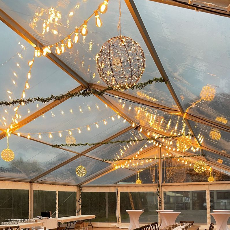 Tente événementielle transparente décorée de guirlandes lumineuses et de boules suspendues.