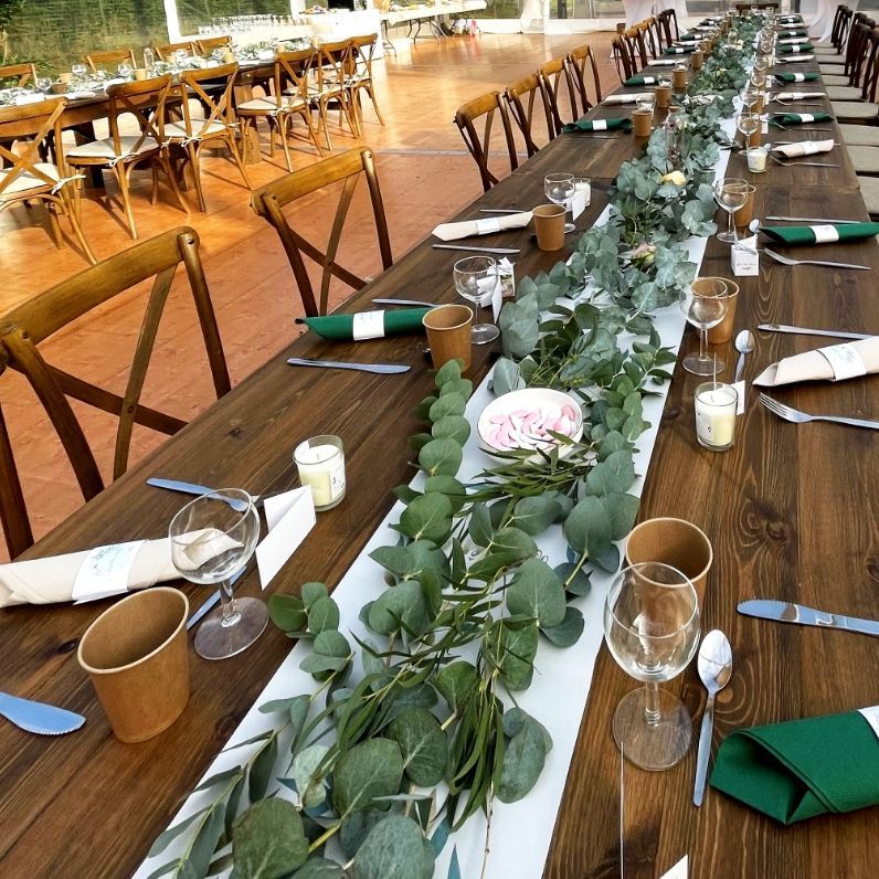 De longues tables en bois sont dressées pour une réception, ornées de guirlandes d'eucalyptus, de vaisselle et de chaises en bois.