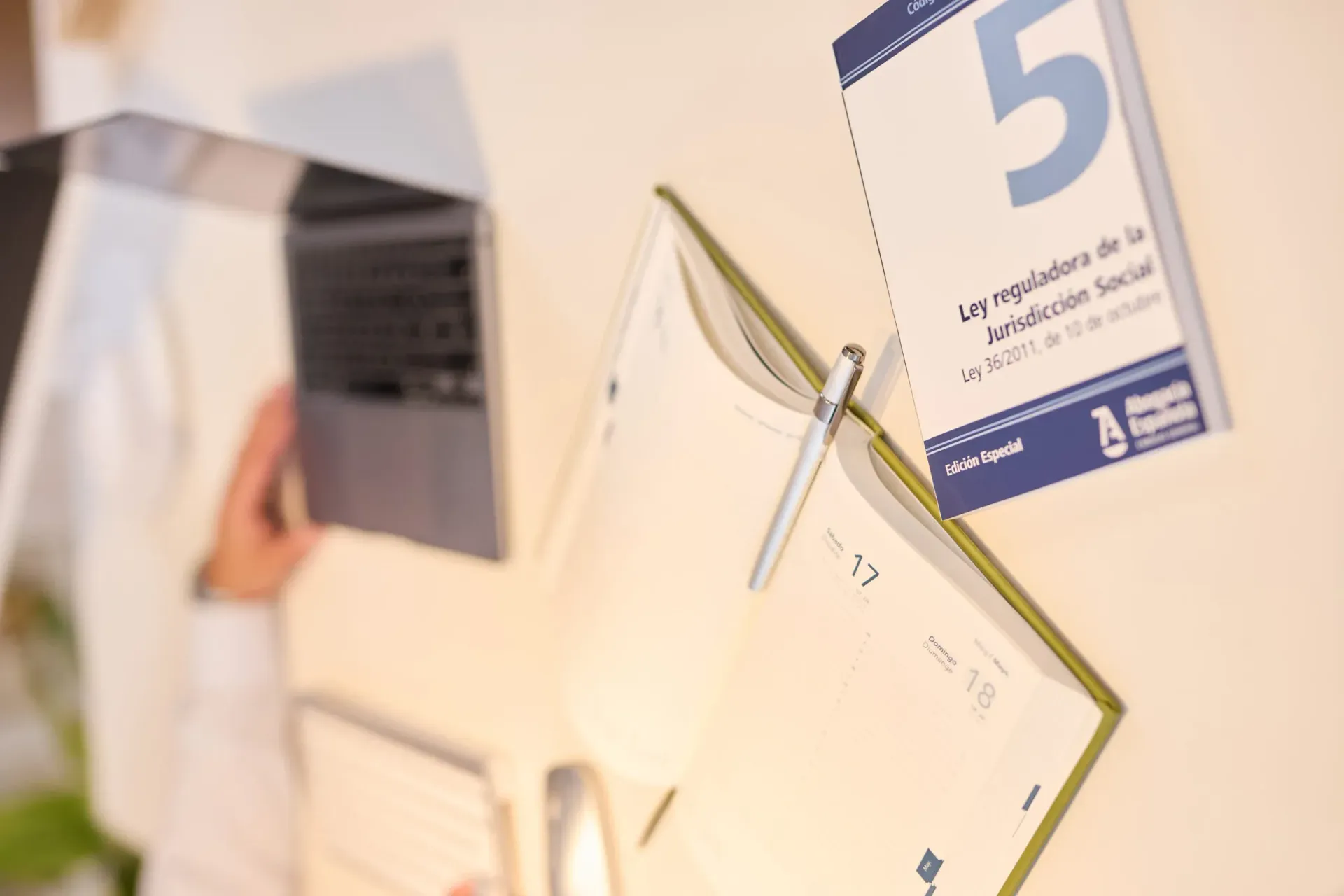 Cuaderno abierto, bolígrafo y calendario sobre un escritorio blanco; persona trabajando en una computadora portátil en el fondo.