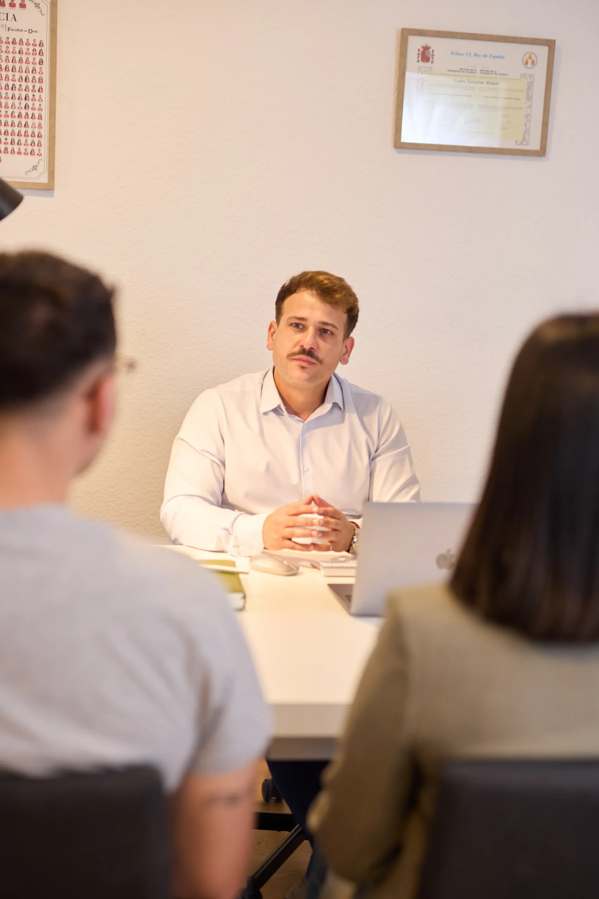 Un hombre está sentado en un escritorio de una oficina, hablando con dos personas sentadas frente a él durante una reunión profesional.