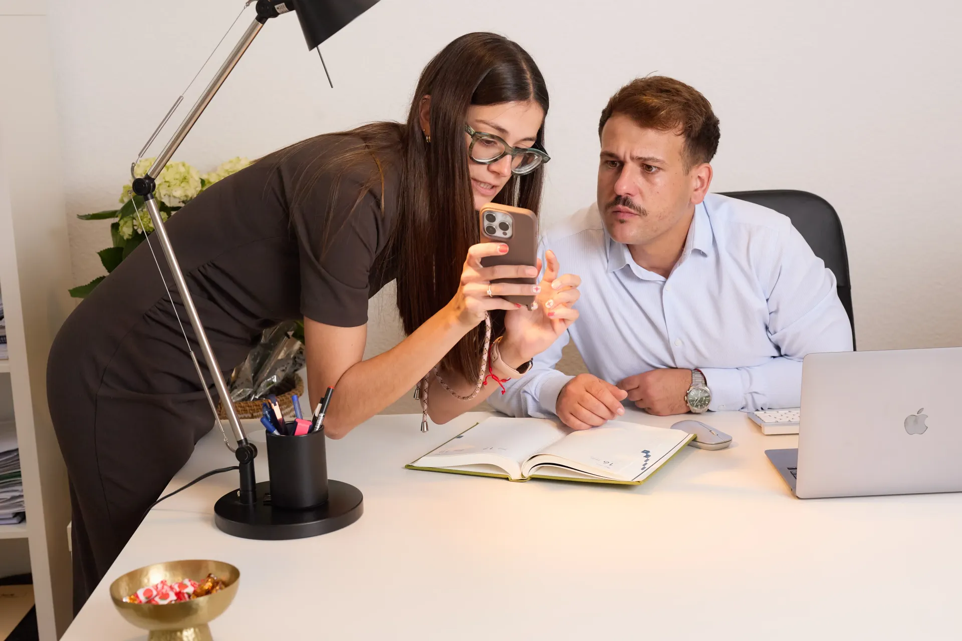 Dos compañeros se inclinan sobre un escritorio blanco, comentando la información que aparece en la pantalla de un teléfono inteligente en una oficina luminosa.