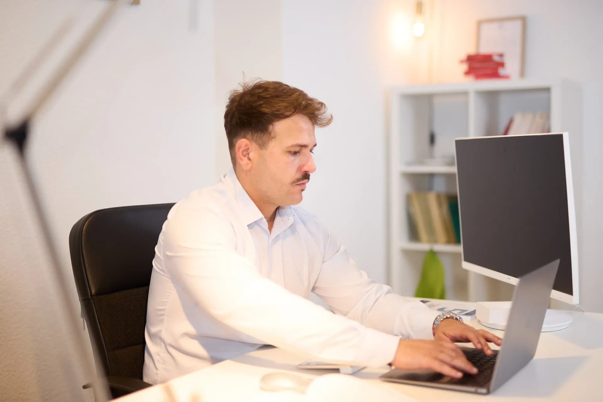 Una persona con una camisa clara está sentada en un escritorio tecleando en una computadora portátil, con un monitor de escritorio y estanterías blancas al fondo.