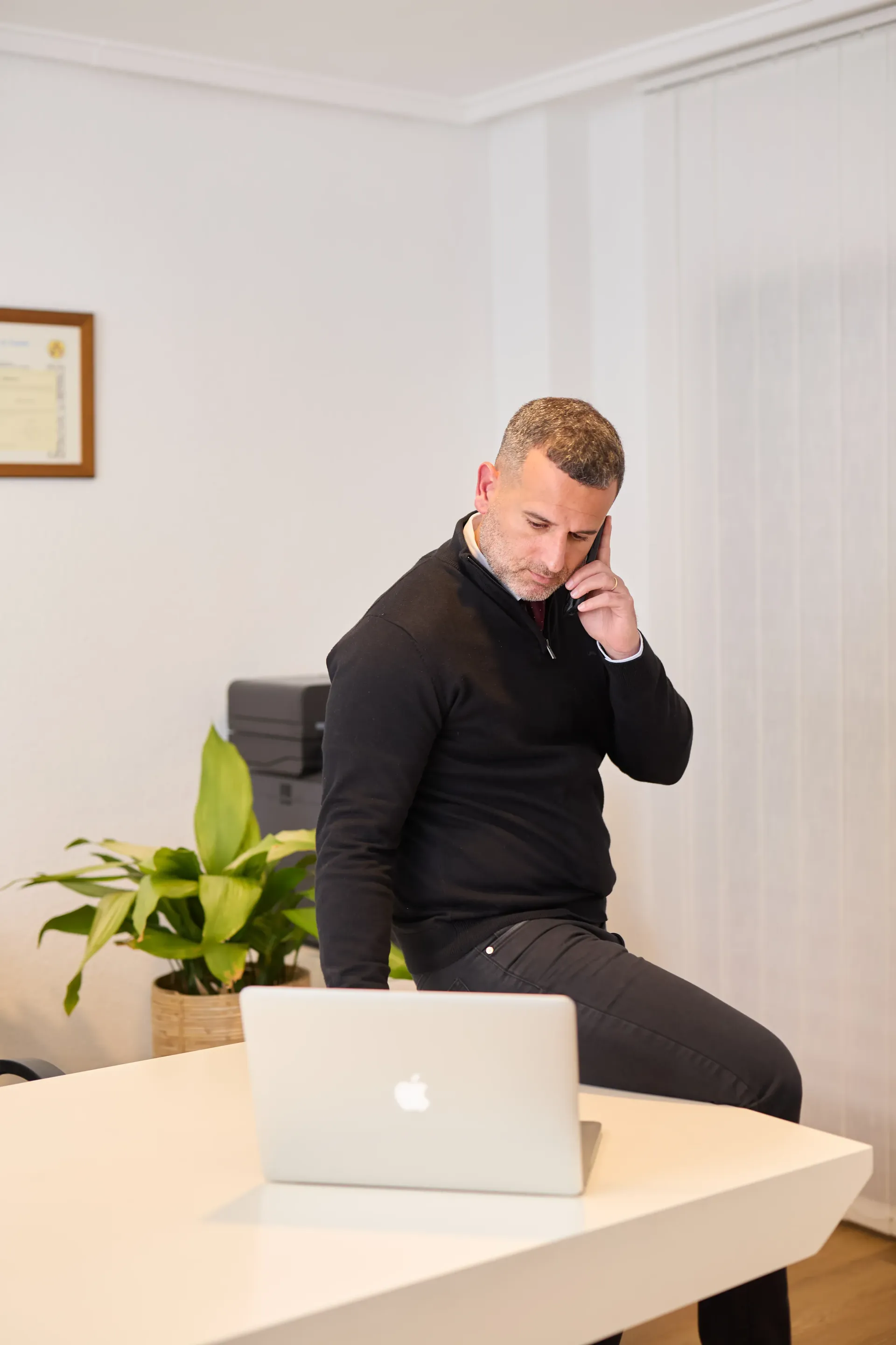 Una persona con un suéter negro está sentada en un escritorio blanco, hablando por teléfono mientras mira una computadora portátil abierta.