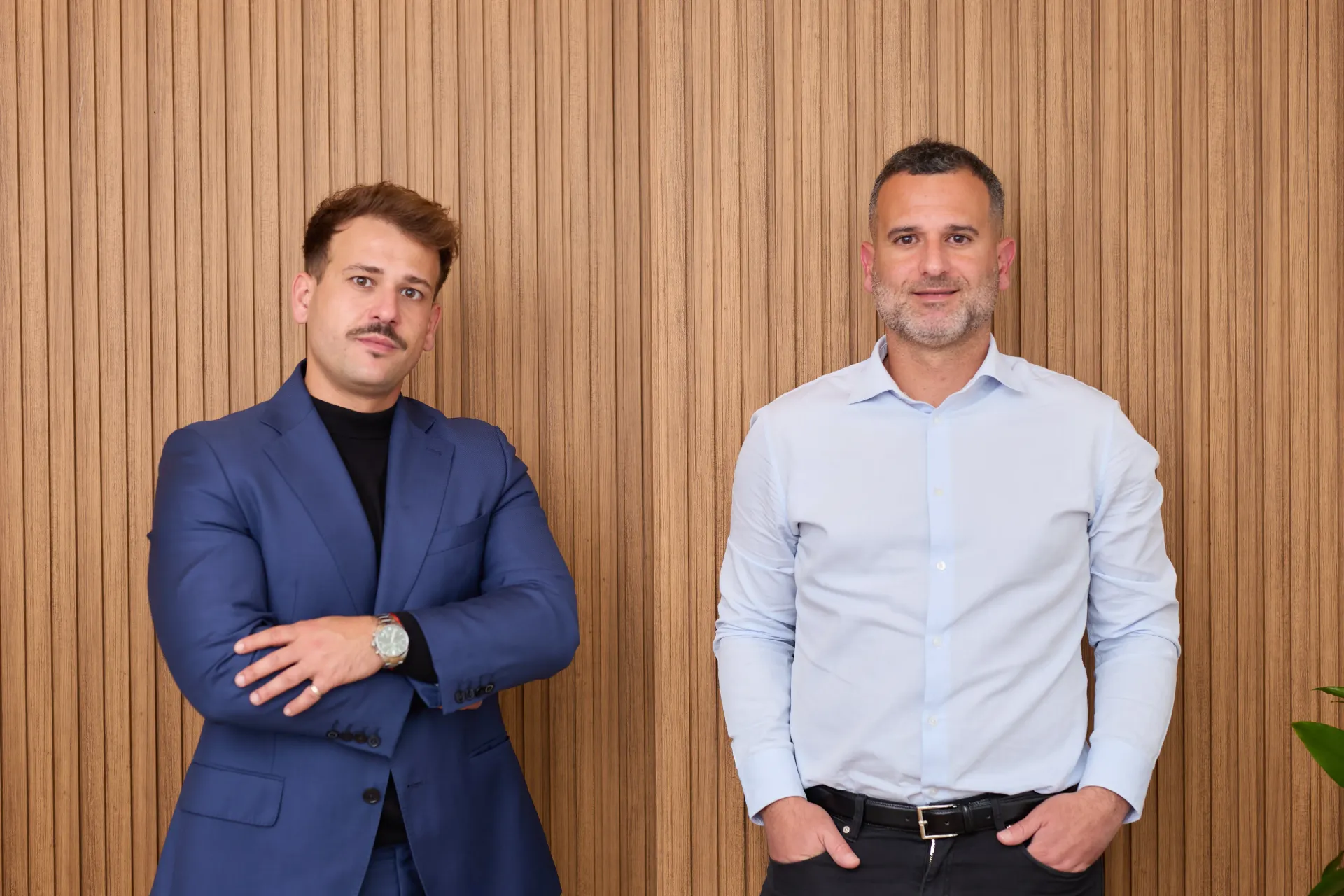 Dos hombres están de pie contra una pared de madera. Uno lleva un traje azul y los brazos cruzados. El otro, una camisa azul claro y las manos en los bolsillos.
