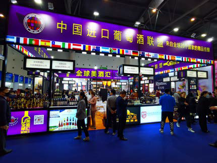 Stand abarrotado en la feria de vinos de China, con coloridas pancartas, luces brillantes y varias personas.