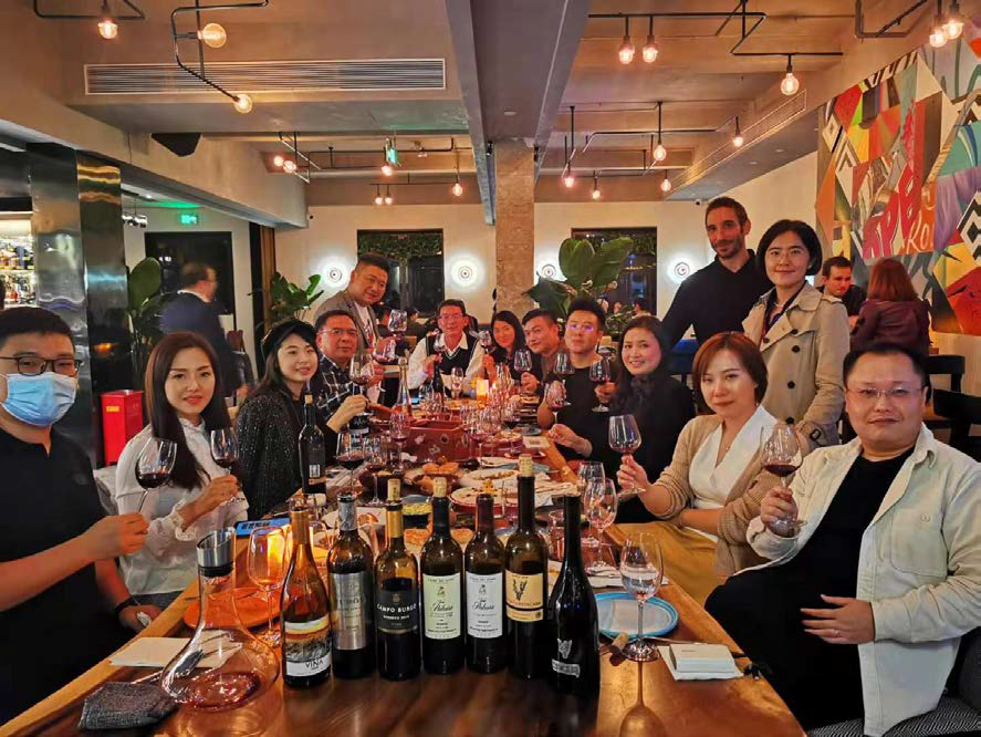 Un grupo de personas brinda alrededor de una larga mesa en un restaurante con iluminación cálida, con botellas de vino.