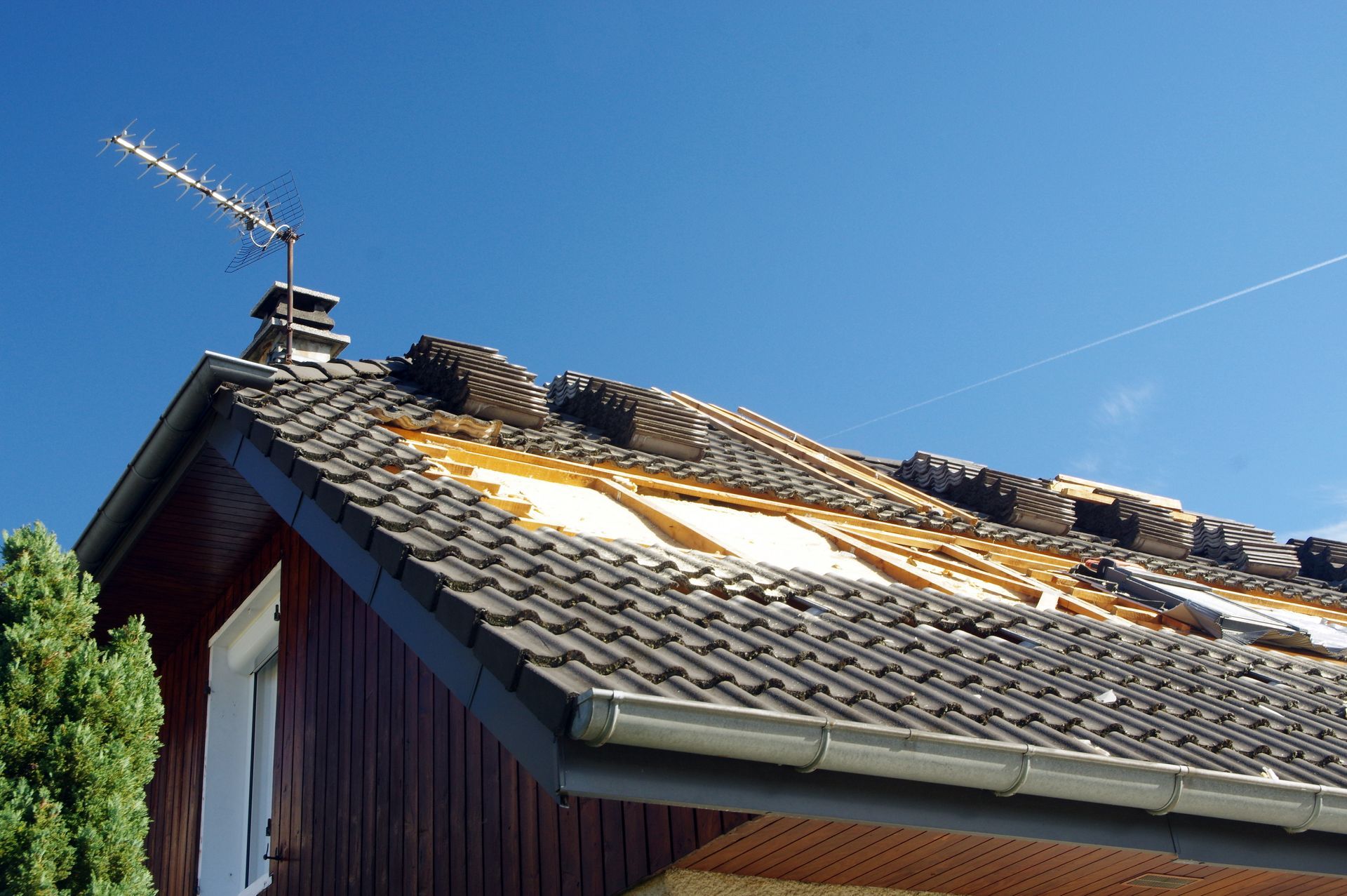 Toiture en cours de pose sous un ciel d'un bleu éclatant, avec une antenne de télévision et un arbre visibles.