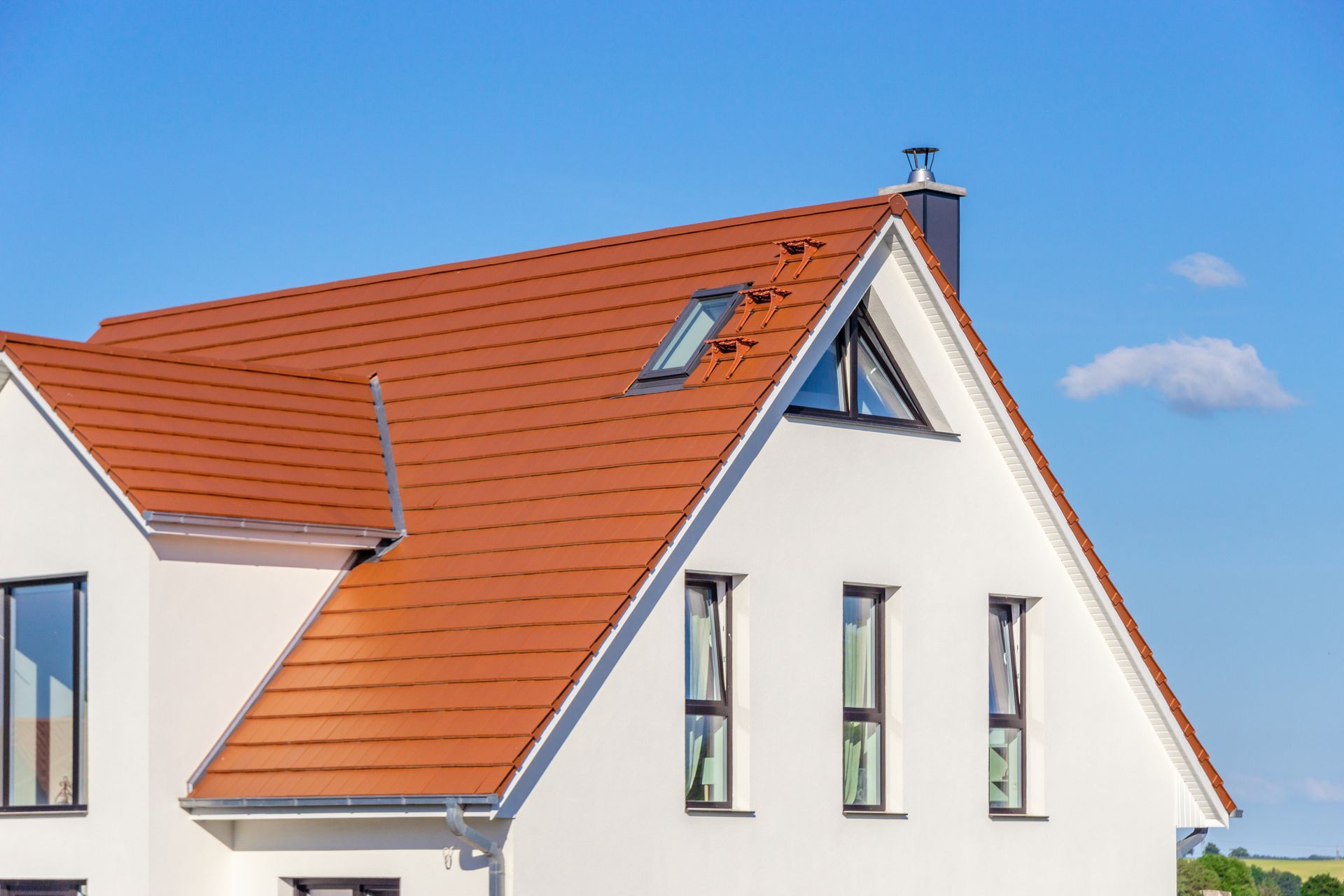 Maison blanche au toit pentu de tuiles rouges avec fenêtres et puits de lumière, se détachant sur un ciel bleu.