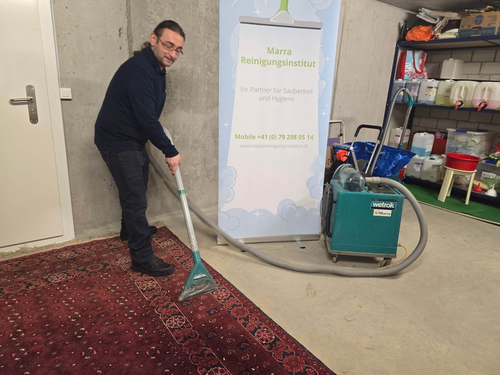Mann saugt drinnen einen roten Teppich mit einem Staubsauger; im Hintergrund ist ein Schild zu sehen.