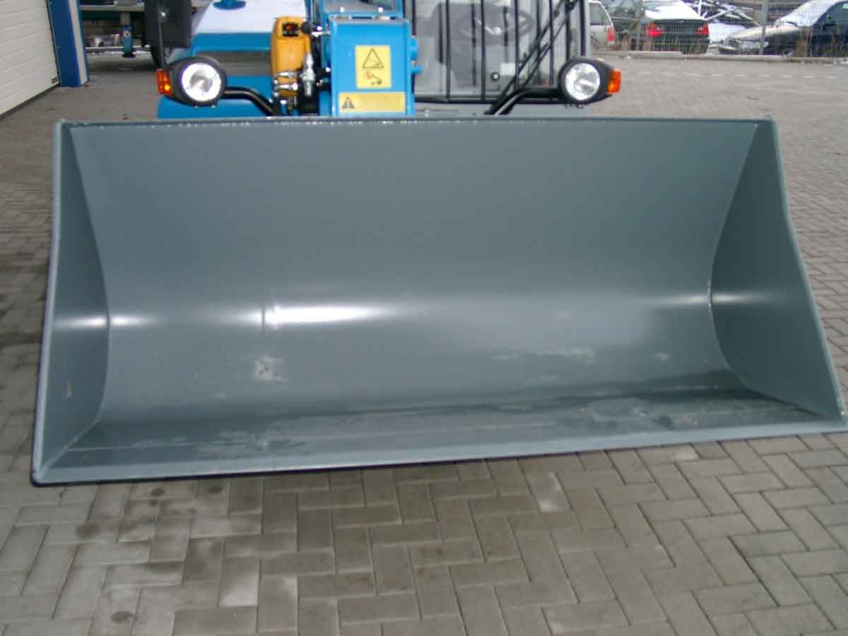 Großer grauer Industrie-Schaufelaufsatz auf einer gepflasterten Fläche, im Hintergrund Maschinen.