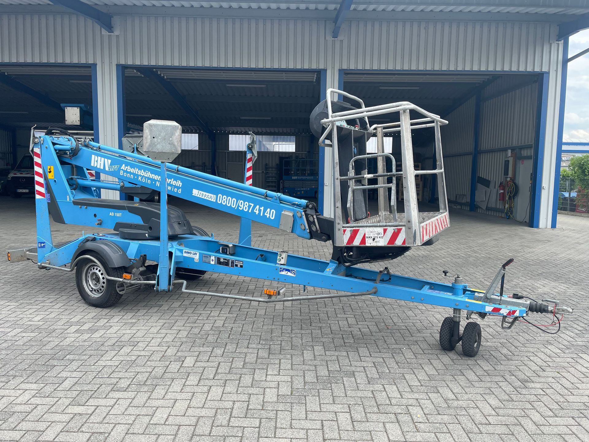 Eine blaue, fahrbare Hubarbeitsbühne mit eingeklapptem Arm und montiertem Korb steht vor einem Lagerhaus.