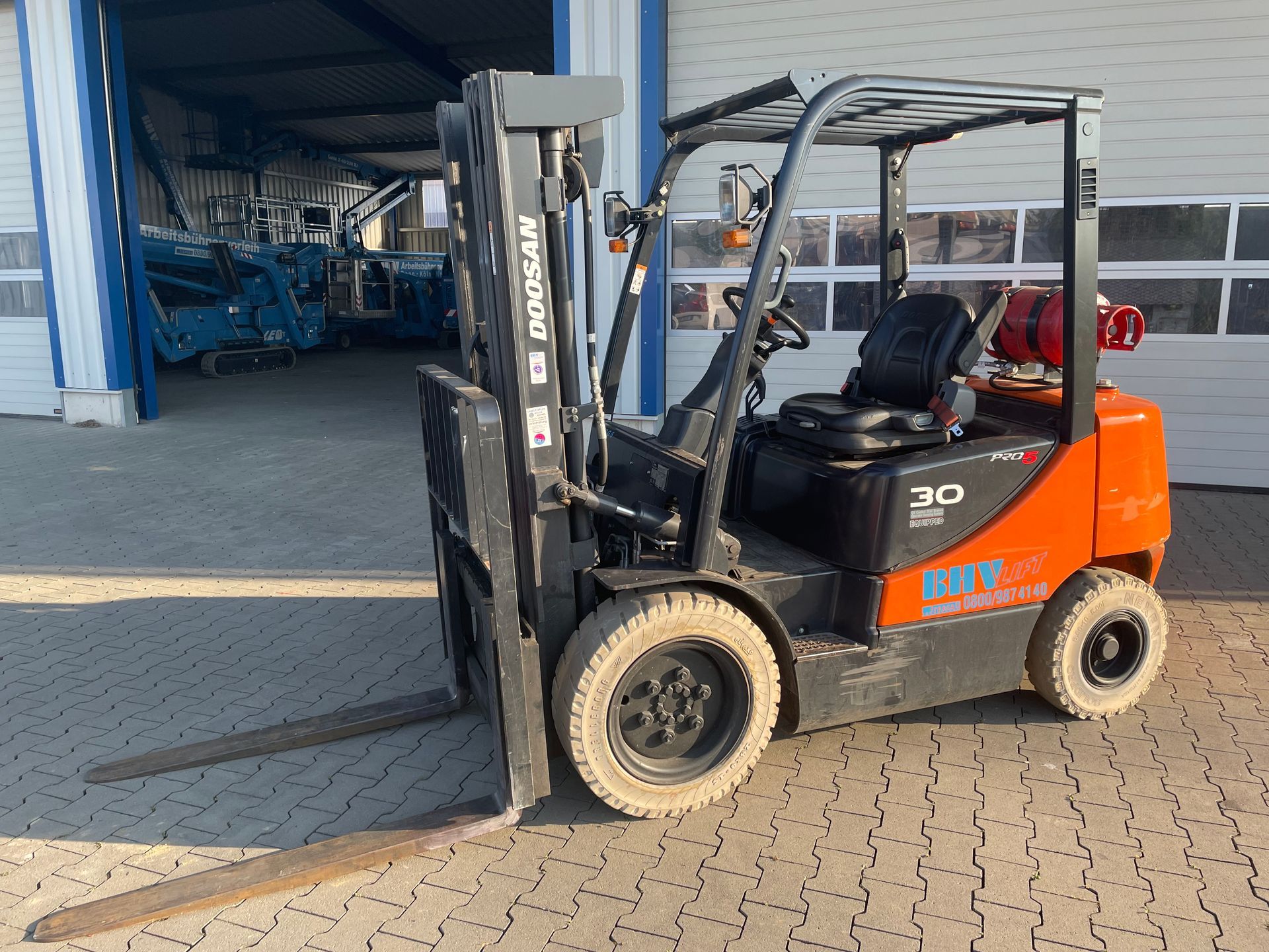 Ein orangefarbener Gabelstapler parkt vor dem Eingang einer Lagerhalle; im Inneren befinden sich blaue Maschinen.