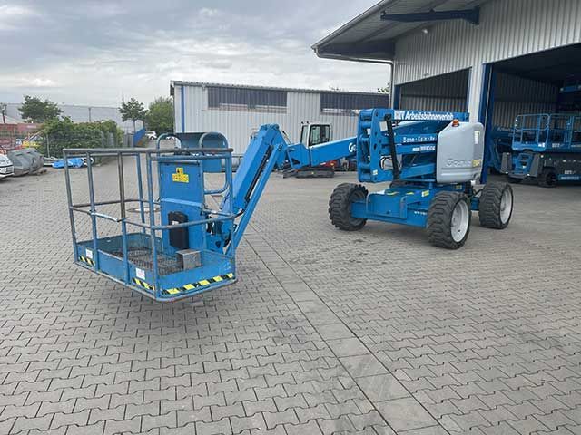Blaue Hubarbeitsbühne mit angehobenem Arbeitskorb, die vor einem Lagerhof geparkt ist.