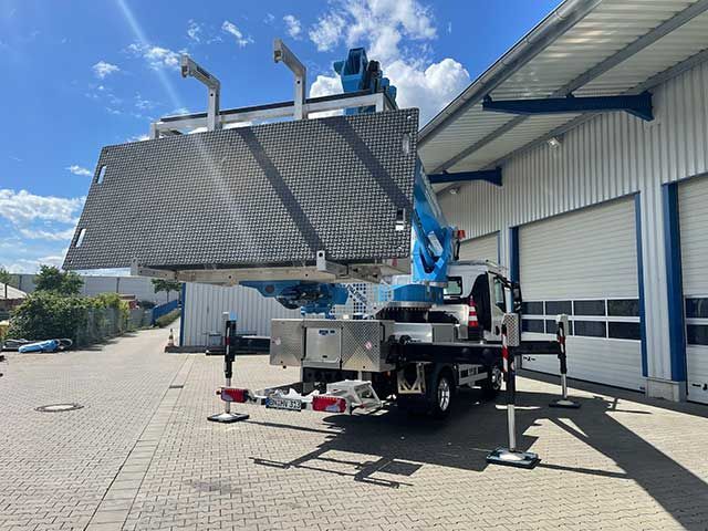 Ein blauer Kranwagen hebt eine große Metallplatte vor einem Industriegebäude an.