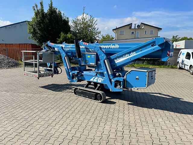 Eine blaue Hubarbeitsbühne steht auf einem gepflasterten Parkplatz neben Gebäuden unter klarem Himmel.