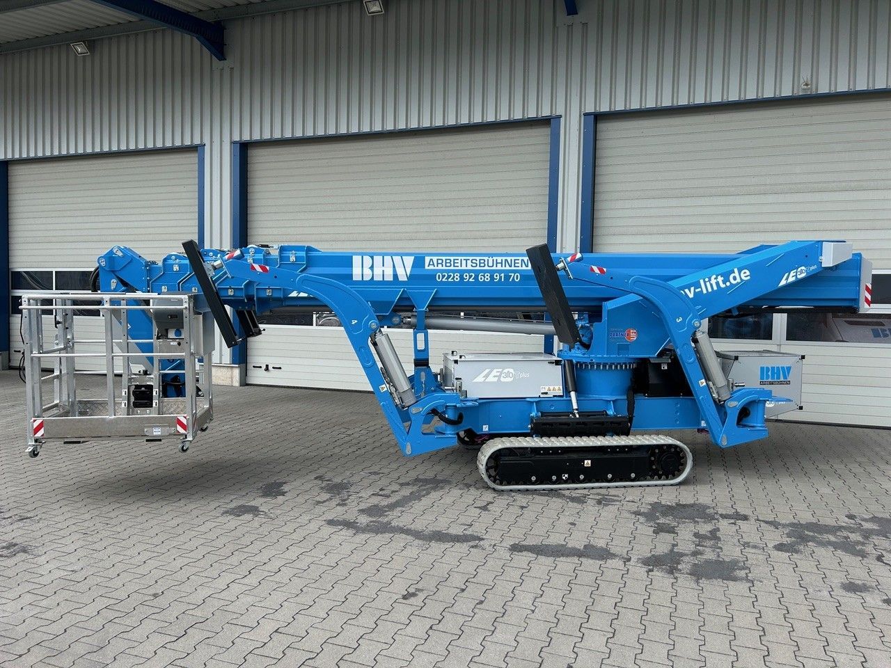 Eine blaue, kettengetriebene Hebebühne ist vor einem Lagerhaus geparkt; daran ist ein kleiner Metallarbeitskorb befestigt.