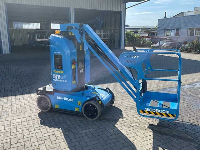 Eine blaue Gelenkteleskoparbeitsbühne mit angehobenem Arbeitskorb und großen Reifen steht vor einem Lagerhaus.